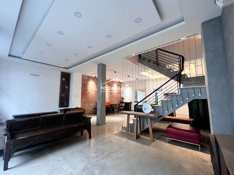 The spacious living area showcases an industrial-chic aesthetic with polished concrete floors, a striking exposed brick feature wall, and a central structural column. A floating staircase with a minimalist metal railing serves as a focal point, connecting the open-plan ground floor to the upper levels while maintaining an airy, unobstructed flow.