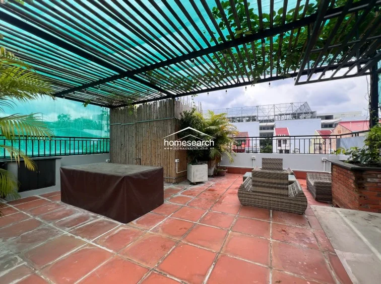 This expansive rooftop terrace is partially covered by a slatted pergola with green netting, offering a shaded space for outdoor dining or lounging. The area is equipped with a large covered table, wicker outdoor furniture, and a built-in cabinet, all set on terracotta-tiled flooring with a perimeter of potted greenery.