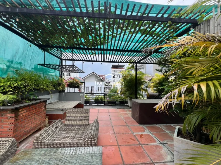This expansive rooftop terrace features a shaded pergola with integrated string lighting, perfect for outdoor relaxation or entertaining. The space is well-maintained, offering comfortable lounge furniture, lush potted greenery, and a brick-built planter box, all set against a backdrop of urban residential views.