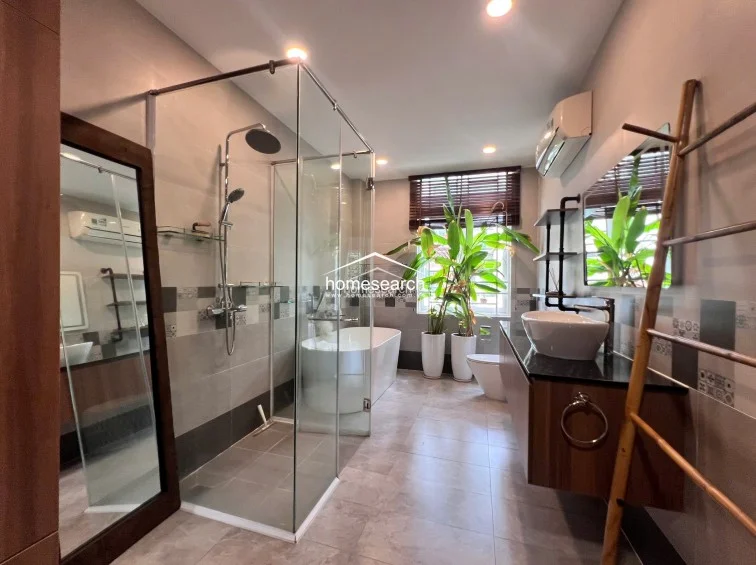 This luxurious, spa-like bathroom features a freestanding soaking tub, a separate glass-enclosed shower, and a floating vanity with a vessel sink. The room is brightened by a large window with wooden blinds and includes thoughtful touches like a decorative ladder rack and an AC unit for climate control.