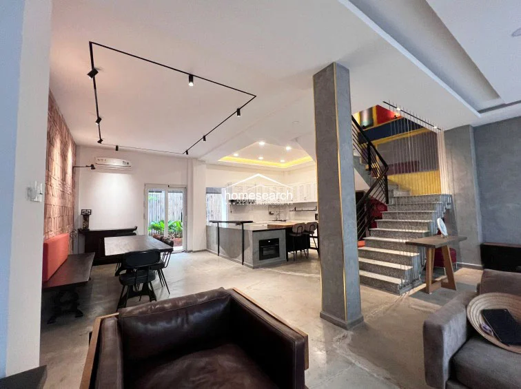 This wide-angle view highlights the cohesive open-plan layout, where the living, dining, and kitchen areas blend into one functional space. The design utilizes track lighting for a contemporary feel, complemented by the warm textures of the brick wall and the clean lines of the concrete flooring and staircase.