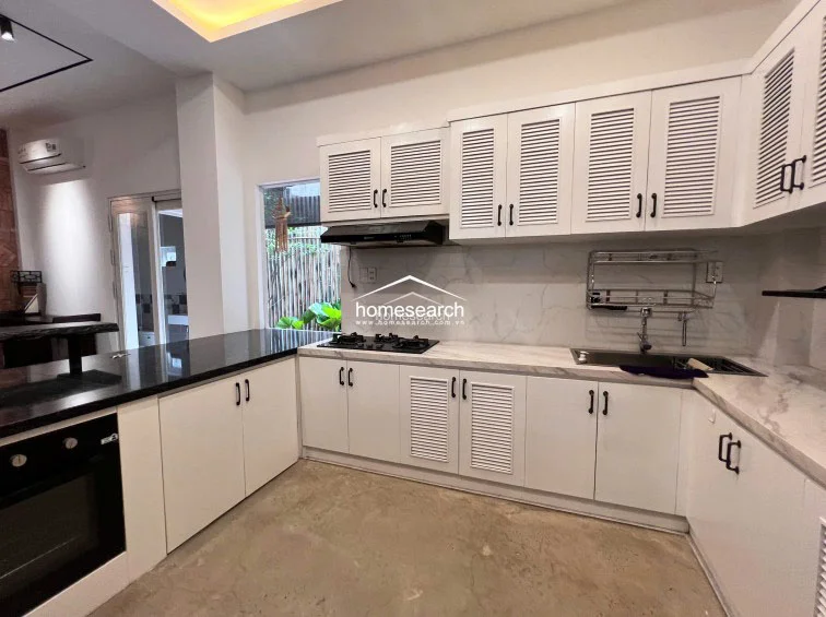The kitchen is highly functional, featuring ample storage within the white louvered cabinets and a spacious marble-topped workspace. It includes essential modern equipment such as a gas cooktop, range hood, and a built-in oven, all set against a clean, neutral backdrop that maximizes the sense of light and space.
