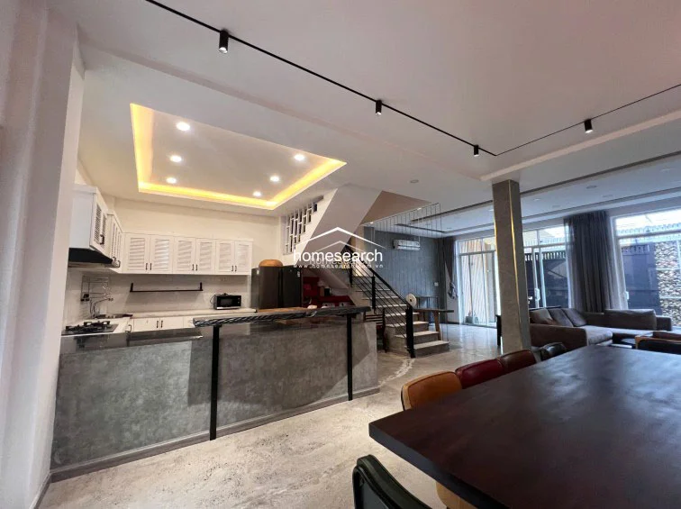 This open-concept living and dining area features a modern kitchen with a concrete-finish breakfast bar and white louvered cabinetry. The space is illuminated by sleek track lighting and recessed ceiling lights, with a staircase providing a central architectural focal point that leads to the upper levels.