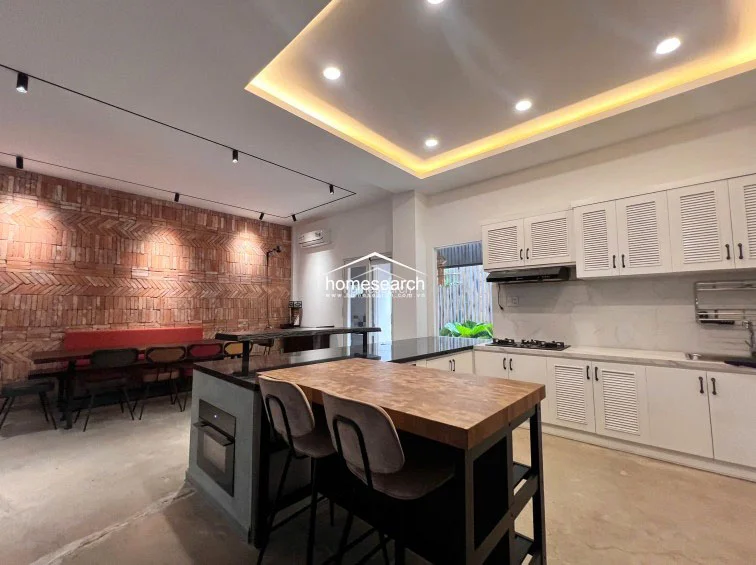 This kitchen area highlights a striking herringbone-patterned brick accent wall that complements the industrial-style breakfast island and wooden countertop. The layout is highly functional, featuring ample white cabinetry, a built-in oven, and a gas cooktop, all set against a neutral, well-lit backdrop.
