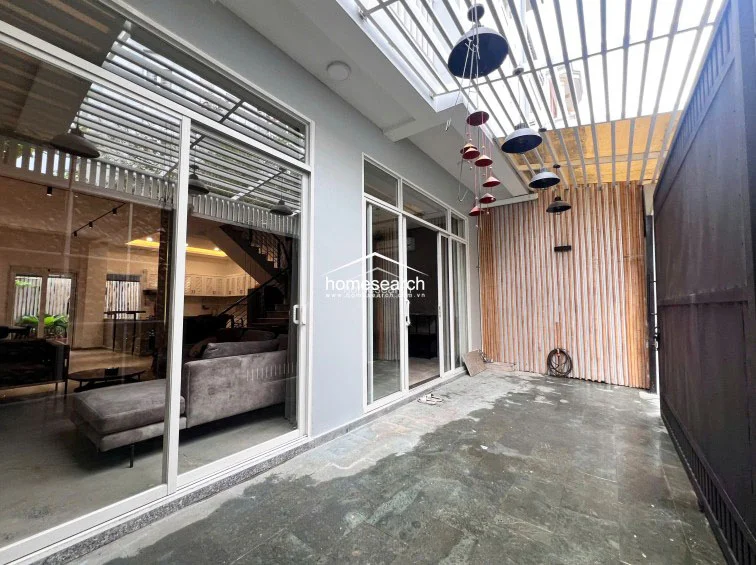 This covered outdoor patio serves as a seamless transition between the indoor living area and the exterior, featuring large sliding glass doors. The space is decorated with hanging lanterns and bamboo-style wall paneling, creating a warm, inviting atmosphere on the stone-tiled floor.
