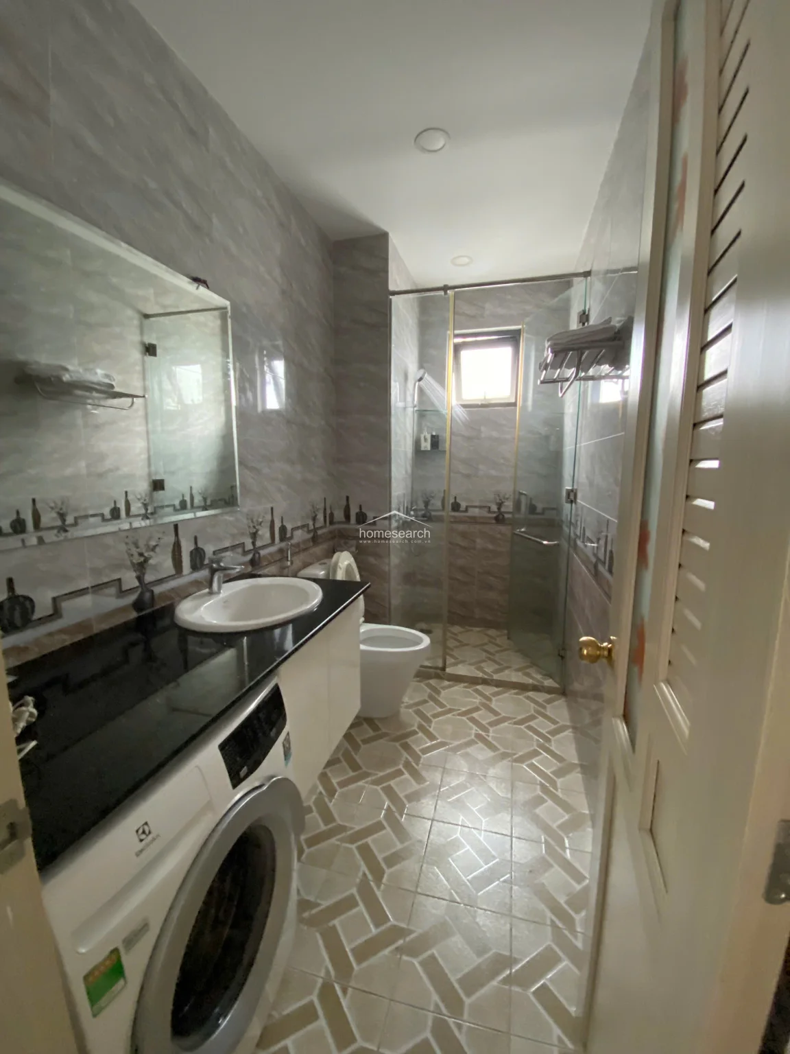 This bathroom features a sophisticated layout with a black granite countertop vanity, an integrated washing machine, and patterned geometric floor tiles. The walk-in shower is enclosed by a glass partition, and the walls are fully tiled with a decorative border, reflecting a high level of maintenance and modern utility. The space is well-lit with recessed ceiling lights and includes a large mirror, maximizing the sense of space in this functional area.