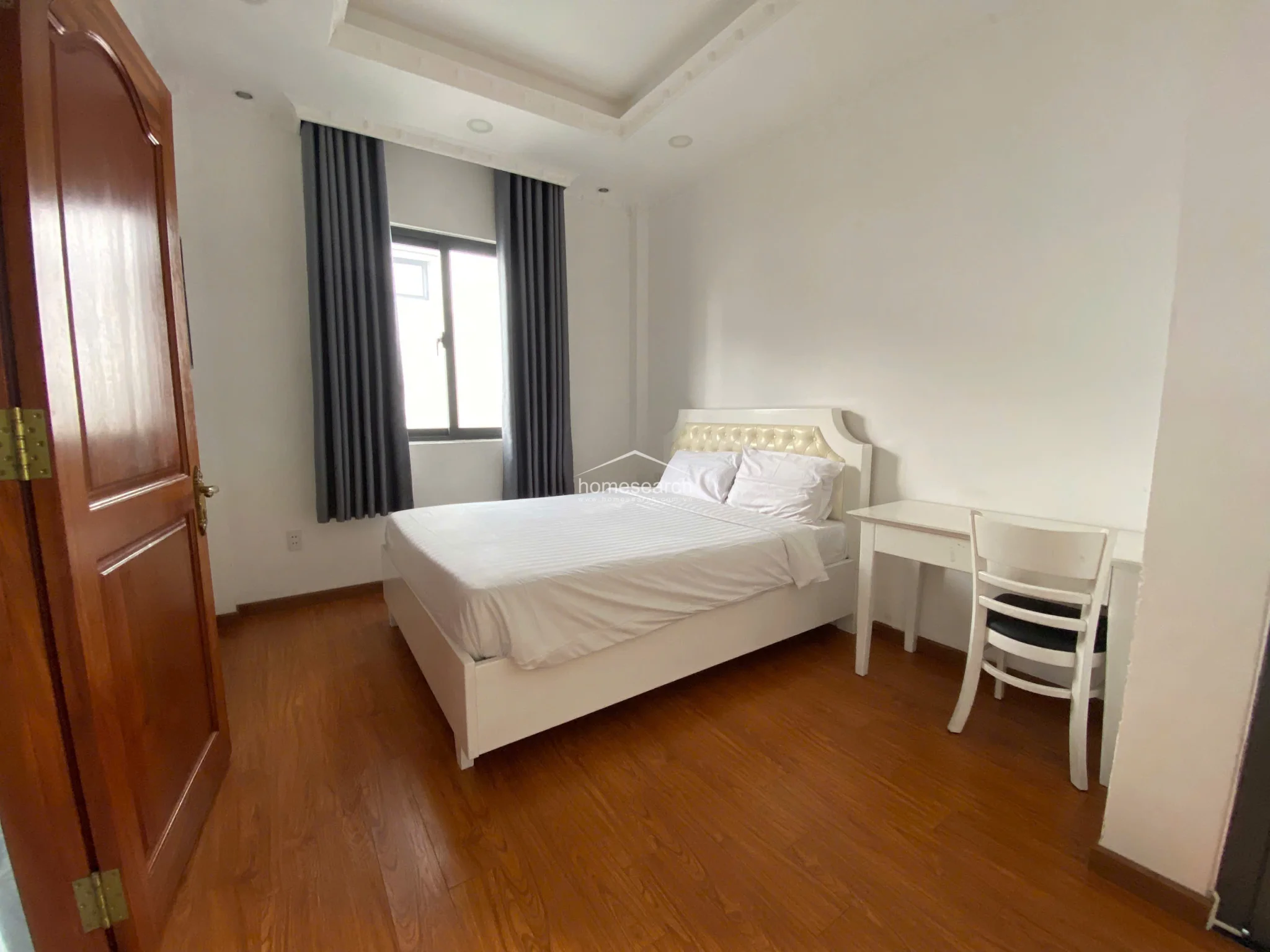 This bedroom is finished with warm wood-tone flooring and crisp white walls, offering a cozy and well-maintained environment. It is furnished with a white bed featuring a tufted headboard and a matching small desk, complemented by dark grey curtains for light control. The room is accessed via a sturdy wooden door and maintains a minimalist aesthetic with high-quality finishes.