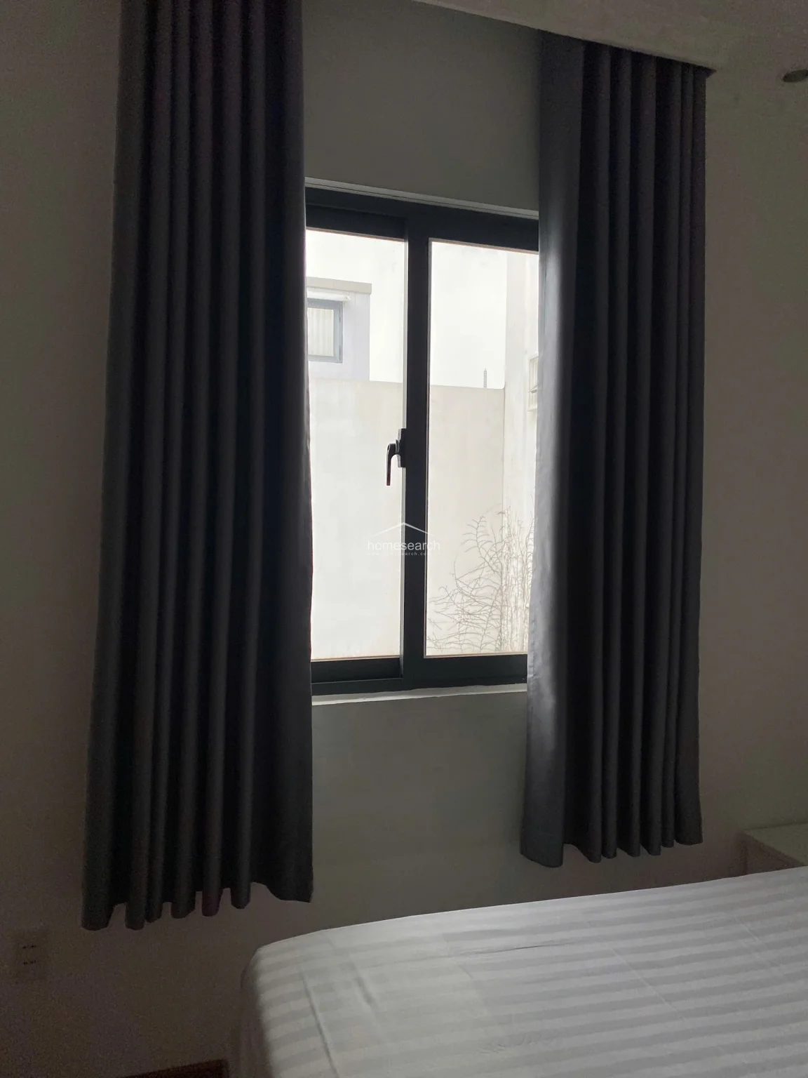 This close-up view highlights a functional window with dark grey curtains, providing natural light and a view of the adjacent building exterior. The window frame is dark-toned, contrasting against the neutral white wall, and the area appears clean and well-maintained. The perspective confirms the room's simple, modern design and focus on essential, practical features.