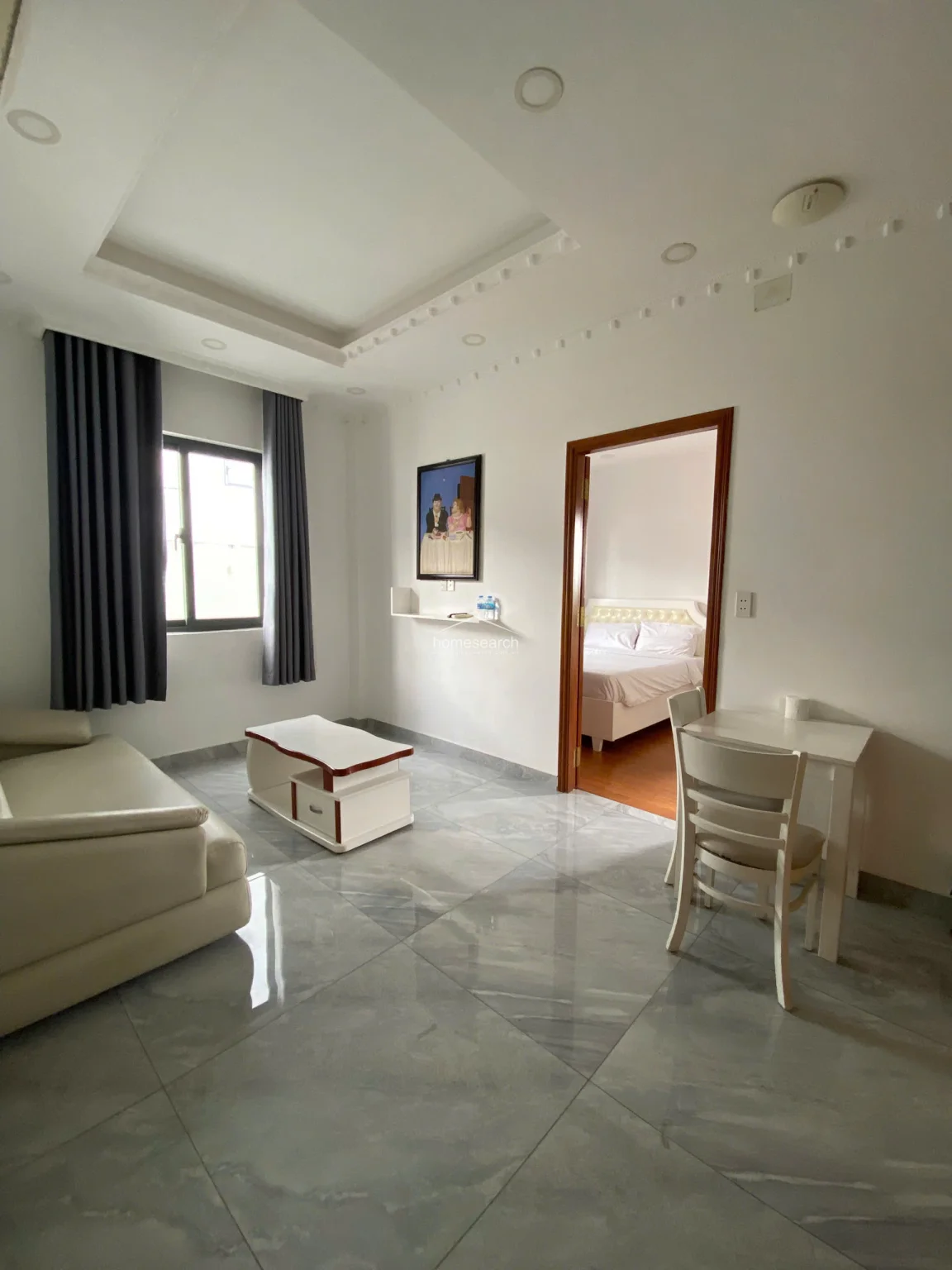 This living area features polished grey marble-style tile flooring and a clean white color scheme, creating a bright and modern atmosphere. The space includes a minimalist coffee table and a small dining set, with a direct, open-flow connection to the bedroom through a wooden-framed doorway. A single window with dark grey curtains provides natural light, while recessed ceiling lighting and a decorative tray ceiling add architectural interest.