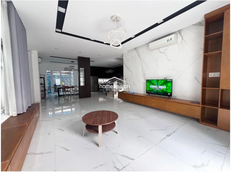This spacious living room features glossy white marble-look floor tiles and a minimalist, contemporary aesthetic with a striking black-and-white ceiling design. The room is anchored by a large, wall-mounted wooden media console and matching shelving, while a modern sculptural chandelier serves as a focal point above the seating area.