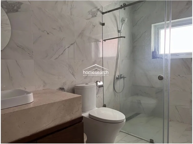 This modern bathroom is finished in high-end, white marble-veined wall and floor tiles, creating a bright and clean atmosphere. It features a glass-enclosed walk-in shower, a sleek wall-mounted toilet, and a vanity with a stone countertop, all appearing to be in excellent, well-maintained condition.