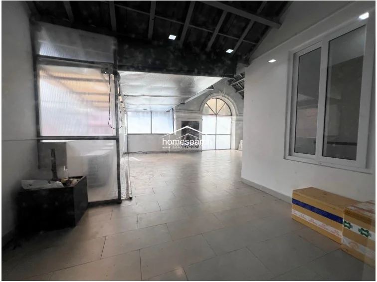 This spacious, open-plan room serves as a versatile living or multi-purpose area with neutral white walls and light-colored tile flooring. The space is illuminated by recessed ceiling lights and features a large, arched window design that provides character and natural light, while a semi-enclosed structure on the left suggests potential for a utility or storage area.