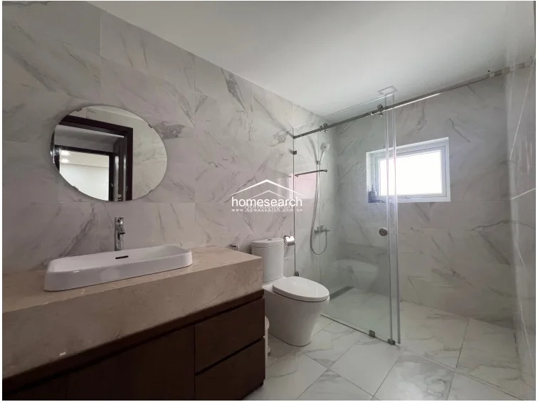 This well-maintained bathroom continues the sophisticated marble-tiled theme, offering a cohesive and luxurious feel. The layout includes a modern wall-mounted toilet, a floating dark wood vanity with a stone countertop, and a glass-enclosed walk-in shower area that maximizes the sense of space.