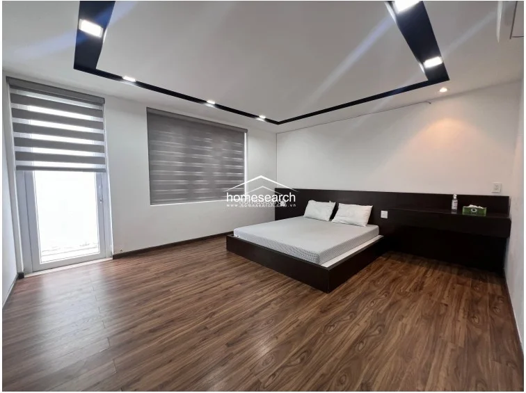 This spacious bedroom features rich wood-look laminate flooring and a striking geometric drop ceiling with integrated recessed lighting. The room is furnished with a low-profile dark wood platform bed and a matching integrated headboard/shelf unit, and it benefits from ample natural light via a large window and a glass door with modern zebra blinds.