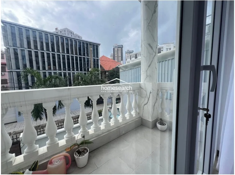 This private balcony offers an outdoor retreat with light-colored floor tiles and classic white balustrades. It is accented by a decorative marble-look pillar and provides an elevated view of the surrounding urban landscape, including nearby modern office buildings.