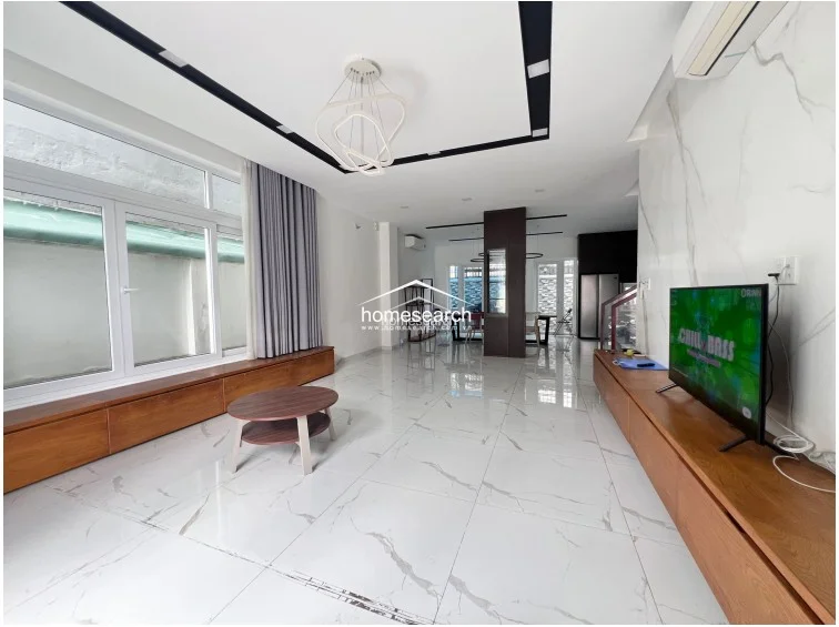 This image provides a wider perspective of the open-plan living area, highlighting the seamless flow from the front entrance through to the dining and kitchen zones. The space is illuminated by large windows with grey curtains and benefits from a wall-mounted AC unit, maintaining a consistent, high-quality finish throughout the floor plan.