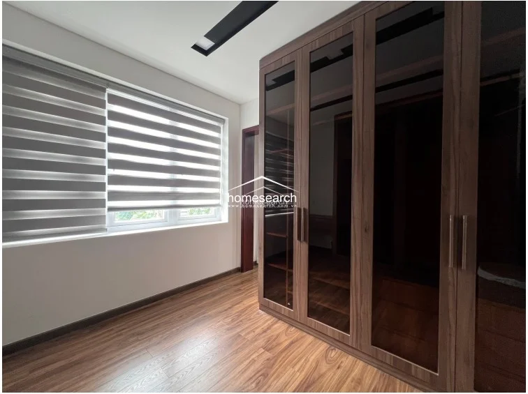 This bedroom space is highlighted by a large window fitted with adjustable zebra blinds, offering flexible control over natural light. The room is equipped with a floor-to-ceiling wardrobe featuring dark glass panels, complementing the rich wood-tone flooring and minimalist white wall finish.