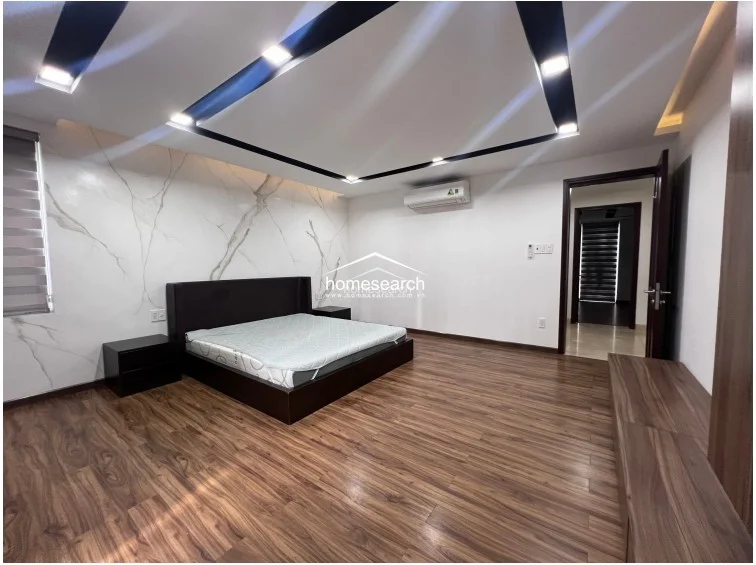 This bedroom highlights a feature wall with elegant marble-patterned cladding behind the dark wood platform bed. The room is climate-controlled with a wall-mounted AC unit and maintains a clean, contemporary aesthetic with minimalist furniture and recessed ceiling lighting.