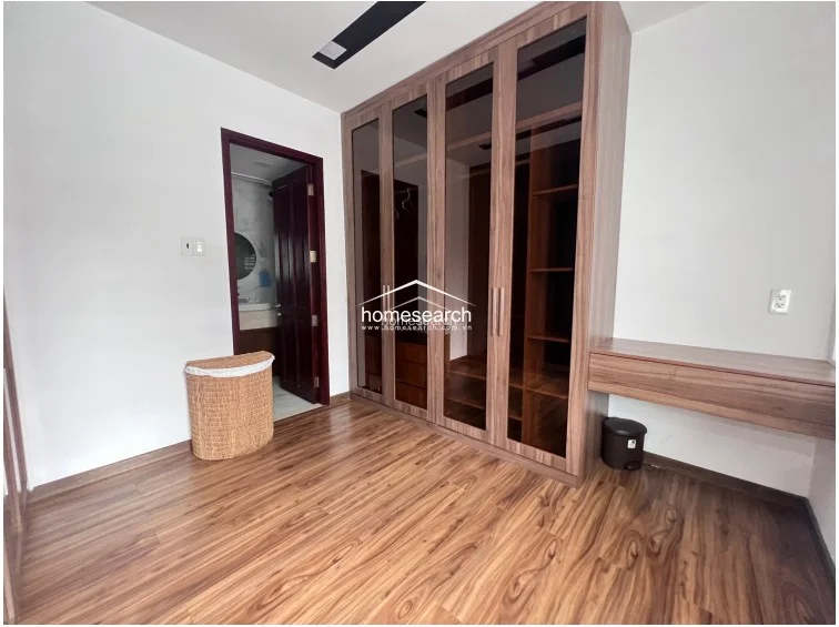 This bedroom features warm wood-look flooring and a large, custom-built wardrobe with dark-tinted glass doors that provide ample storage. The space is finished with clean white walls and includes a built-in desk area, maintaining a modern and well-maintained aesthetic.
