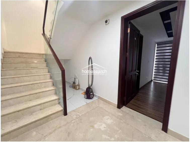 This entryway area showcases a sleek staircase with marble steps and a contemporary glass balustrade with dark wood handrails. A dark wooden door leads into an adjacent room, and the space is kept simple and clean, reflecting a high level of maintenance and modern architectural design.