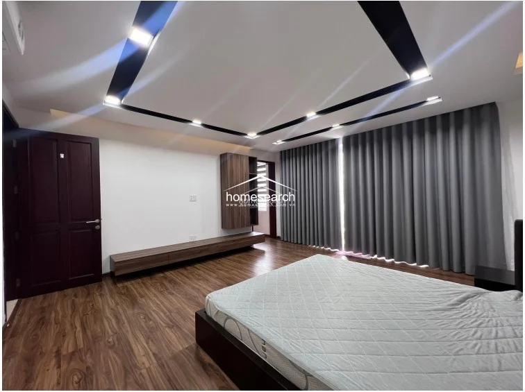 This bedroom is designed with a cohesive dark wood theme, featuring a custom wall-mounted media console and shelving unit. Floor-to-ceiling grey curtains provide full light control, while the room's layout emphasizes a spacious, uncluttered feel with high-quality finishes throughout.