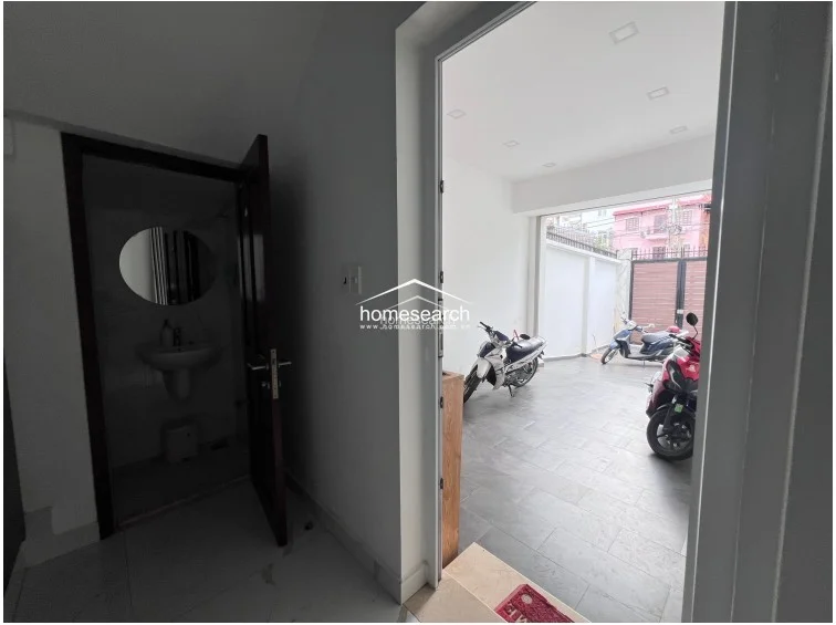 This image shows the ground floor entry, which includes a small powder room and a tiled driveway area currently used for motorcycle parking. The space is utilitarian, featuring a simple pedestal sink in the bathroom and direct access to the main living area through a glass door.