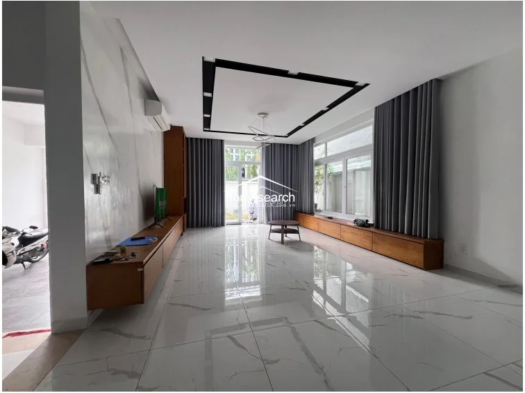 The living room is characterized by expansive white marble-style floor tiles and a striking feature wall with marble cladding. Large windows draped in floor-to-ceiling grey curtains allow for significant natural light, while the recessed ceiling lighting and minimalist decor create a sophisticated, open-plan atmosphere.