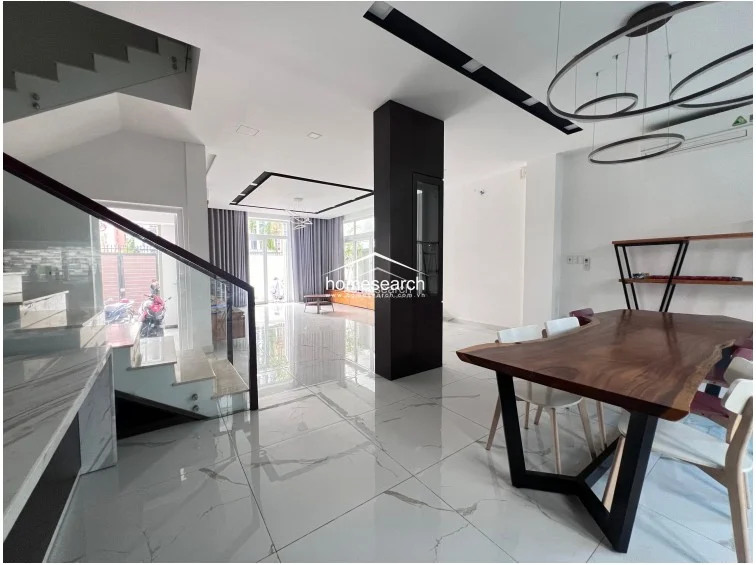 The dining area flows seamlessly into the living space, anchored by a large, natural-edge wooden dining table and modern chairs. A central dark pillar provides a bold architectural contrast, while the open layout and bright, polished flooring emphasize the home's spacious and airy design.