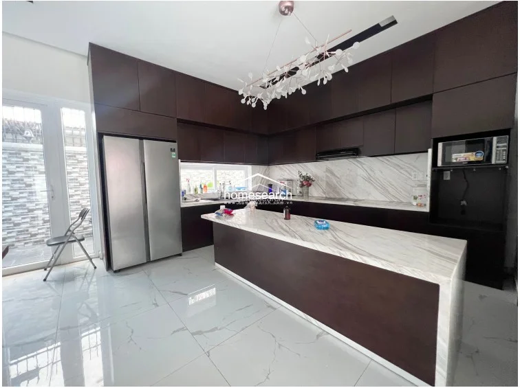 The kitchen is designed with a sophisticated contrast, featuring dark wood cabinetry and a large central island with a waterfall-edge marble countertop. It is well-equipped with a stainless steel side-by-side refrigerator, built-in microwave, and modern pendant lighting, with direct access to an outdoor area through a glass door.