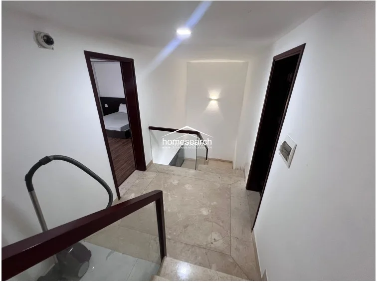 This landing area features elegant marble flooring and a modern glass-railed staircase that connects the levels with an open, airy flow. The space is finished in clean white tones with warm accent lighting and includes a security camera for added safety.