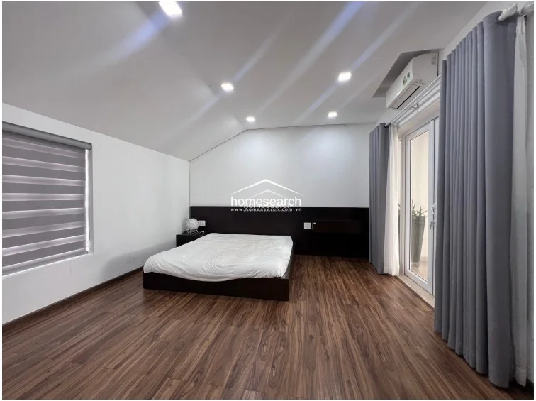 This bedroom features warm wood-look flooring and a sloped ceiling, creating a cozy atmosphere with ample natural light from the large window and glass balcony door. It is equipped with a wall-mounted AC unit, recessed lighting, and a dark wood headboard with integrated bedside shelving.