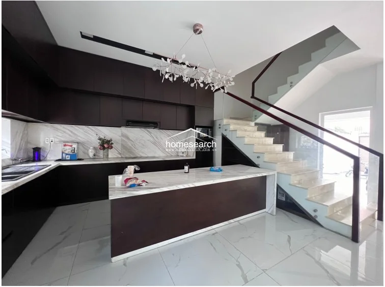 This modern kitchen features dark, floor-to-ceiling cabinetry contrasted by a white marble-veined island and backsplash. The open-plan layout flows seamlessly toward a floating staircase with glass railings, creating a bright and airy atmosphere with high-end finishes.