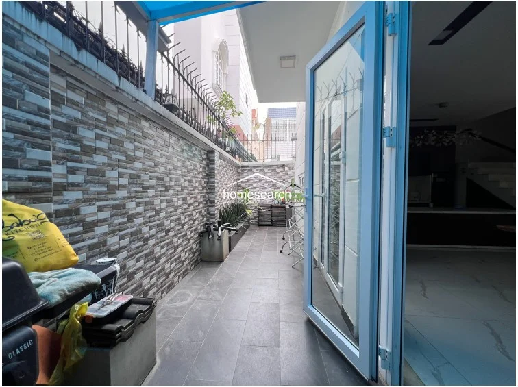 The outdoor side terrace is finished with textured stone-look wall tiles and durable grey floor tiles, providing a functional space for laundry or storage. It is enclosed by a high metal fence for security and connects directly to the interior living space via large glass doors, offering a clean and well-maintained exterior area.