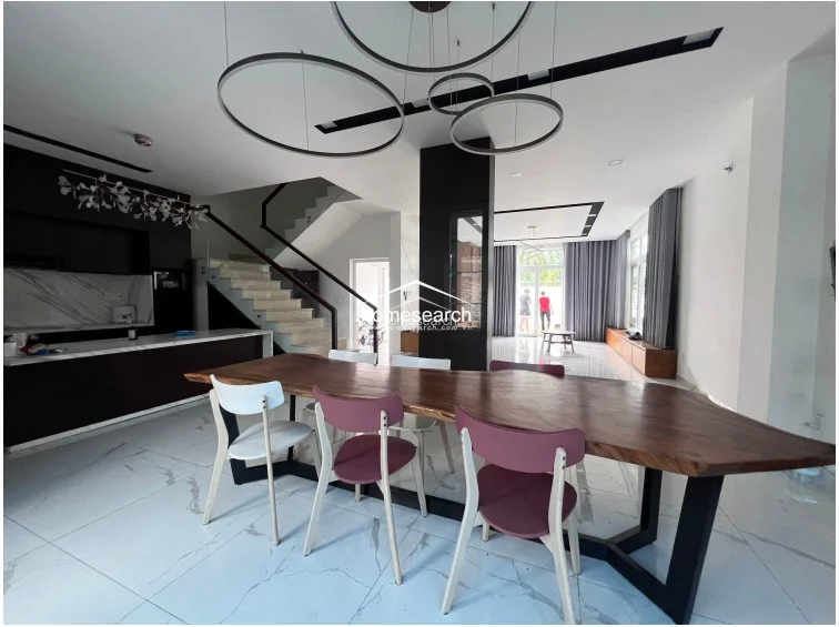 The dining area is anchored by a large, natural-edge wooden table and a striking modern chandelier, serving as a central hub between the kitchen and the living space. The interior maintains a cohesive aesthetic with white marble tile flooring and minimalist, contemporary lighting fixtures throughout.