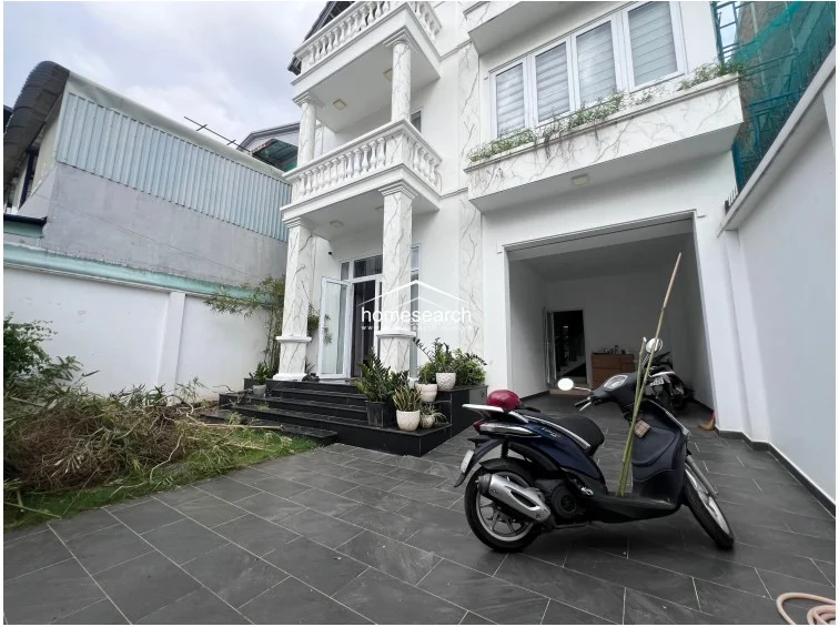 This exterior view showcases a modern multi-story townhouse featuring a covered carport with dark stone tiling and a small front garden area. The architectural design includes prominent white columns with marble-effect detailing, a balcony with balustrades, and large glass doors that provide a clear connection to the interior living space.
