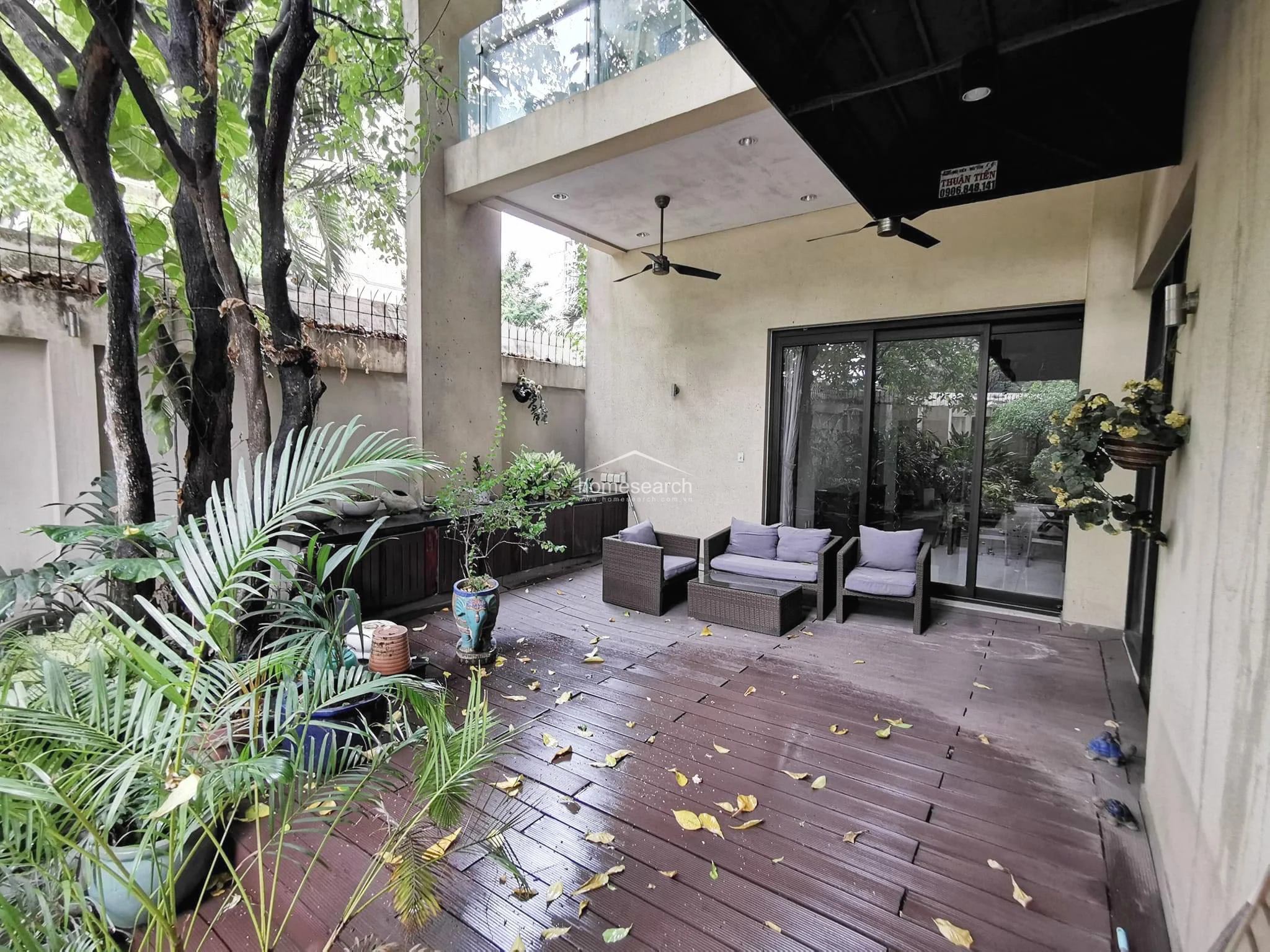 This outdoor terrace features dark-toned composite decking that connects seamlessly to the interior living space through large sliding glass doors. The area is partially covered, equipped with ceiling fans for comfort, and includes a built-in storage console along the wall. While the space offers a relaxing garden view, it shows signs of moderate wear with scattered leaves and damp surfaces.