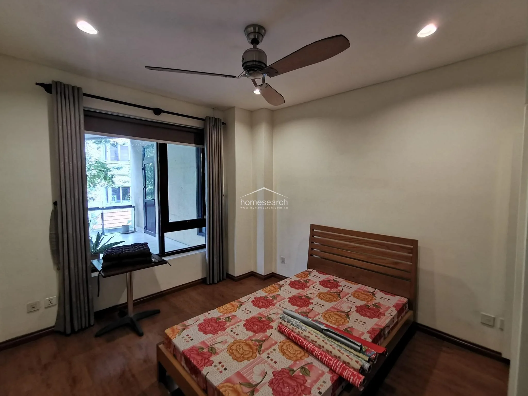 This bedroom features a functional layout with warm wood-look flooring and a wooden bed frame, accented by a floral-patterned bedspread. The room includes a ceiling fan, recessed lighting, and a large window with floor-length curtains that opens onto a private balcony, offering a seamless connection to the outdoors.