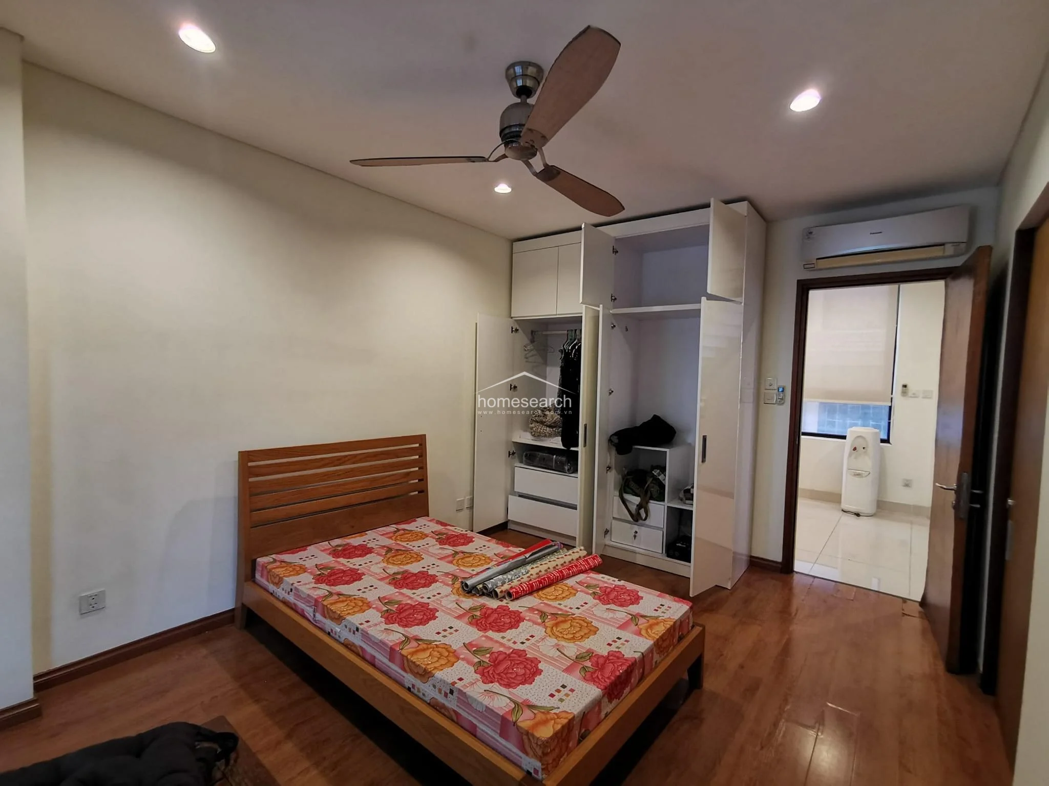 This bedroom features warm wood-tone flooring and a minimalist aesthetic, anchored by a wooden bed frame and a large, white multi-section wardrobe. The room is equipped with a ceiling fan and an AC unit, with a doorway leading to an adjacent tiled space that includes a water dispenser. The overall condition appears well-maintained with clean, neutral wall finishes and recessed lighting.