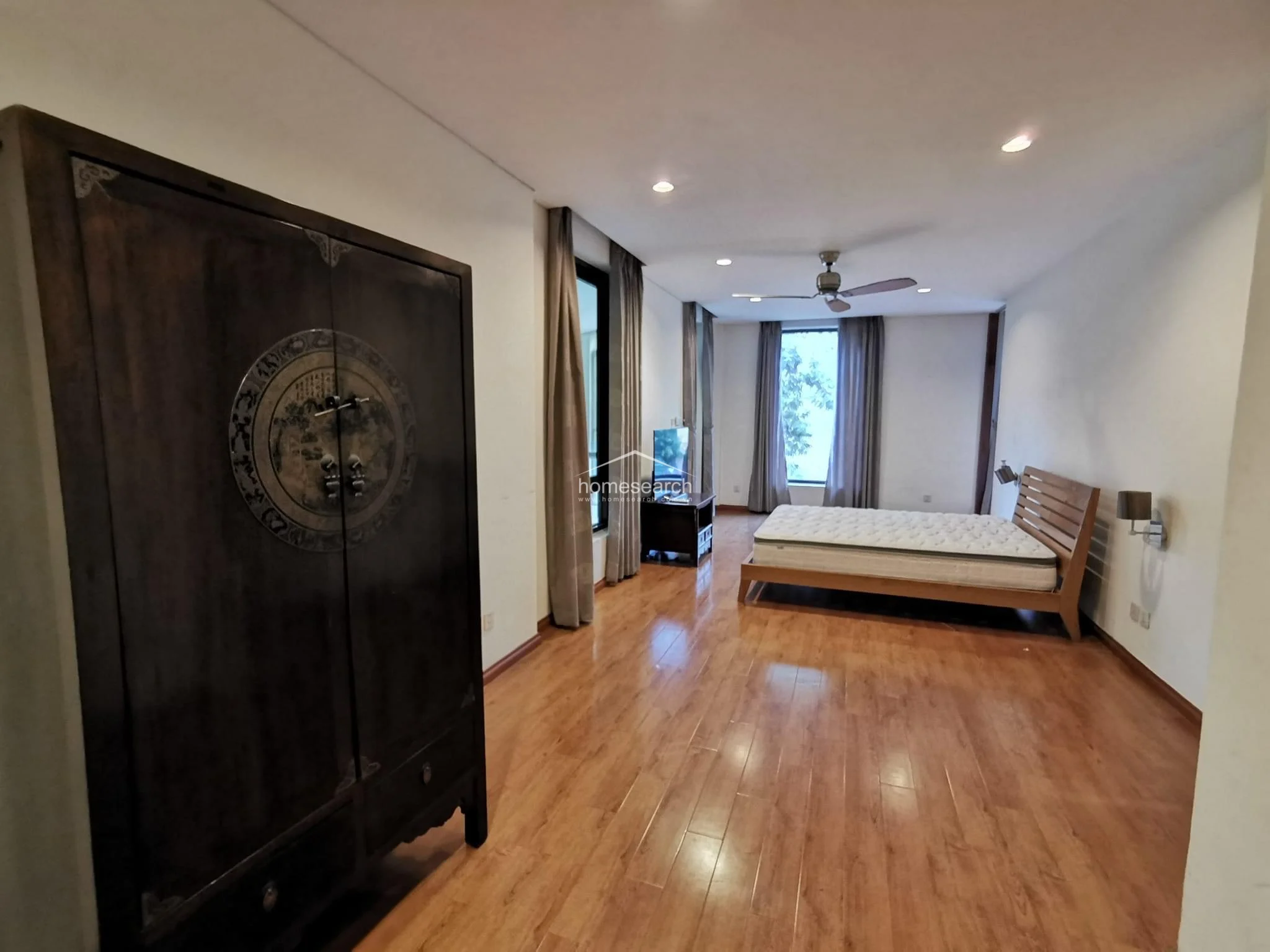This spacious bedroom offers an open-concept layout with polished wood flooring and a large, antique-style dark wood wardrobe that serves as a focal point. The room is bathed in natural light from large windows dressed in floor-to-ceiling curtains, providing a pleasant view of greenery outside. A wooden bed frame and a TV console are positioned to maximize the flow of the room, which is illuminated by modern recessed ceiling lights.