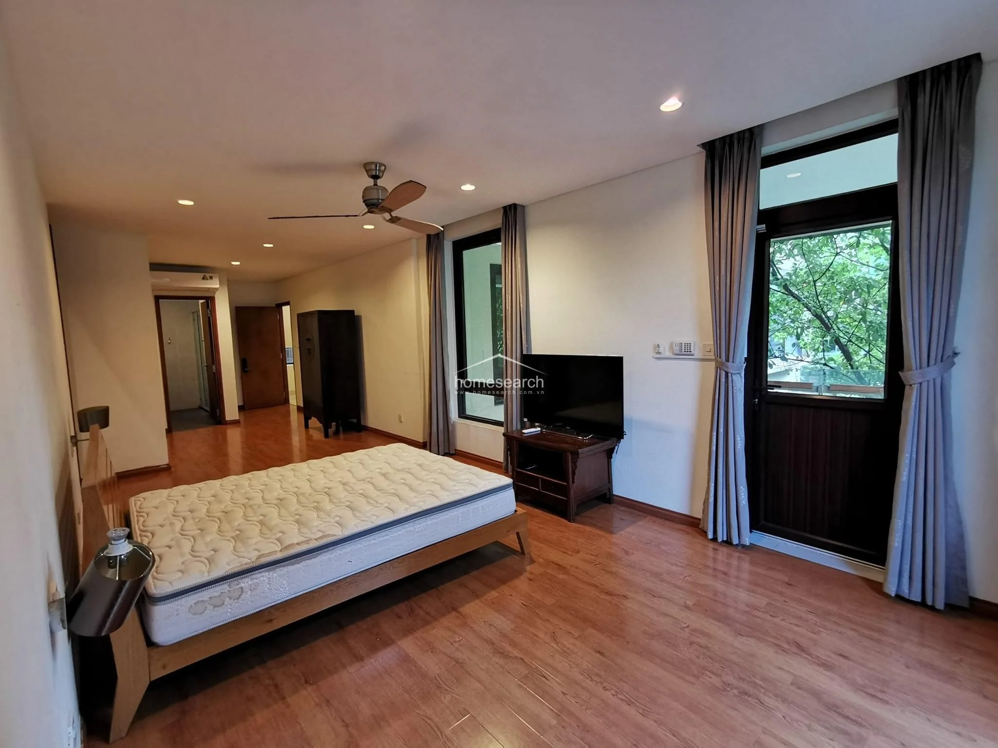 This bedroom continues the theme of wood flooring and neutral walls, featuring a simple bed frame and a television console positioned near the windows. A glass door provides direct access to a small outdoor balcony with lush tree views, enhancing the room's connection to the exterior. The space is uncluttered and well-lit, with a ceiling fan and recessed lighting creating a comfortable, airy atmosphere.