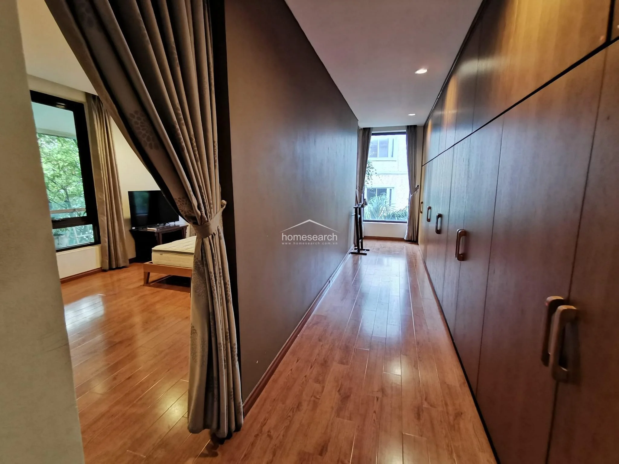 This hallway area acts as a transition space, featuring an extensive wall of floor-to-ceiling wooden cabinetry that provides significant storage capacity. The warm wood flooring extends from the adjacent bedroom, maintaining visual continuity, while a heavy curtain separates the spaces for privacy. A window at the end of the hall invites natural light, making the corridor feel bright and functional.