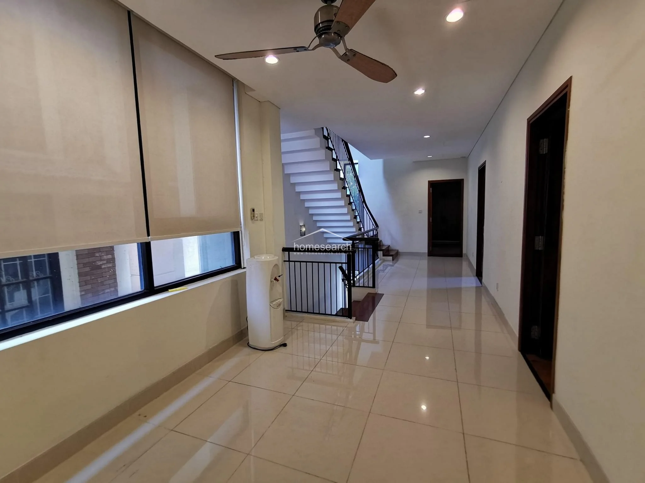 This hallway features polished light-colored tile flooring and a clean, minimalist aesthetic with white walls and recessed ceiling lighting. A central staircase with a black metal railing serves as the focal point, connecting the levels while a ceiling fan provides air circulation.
