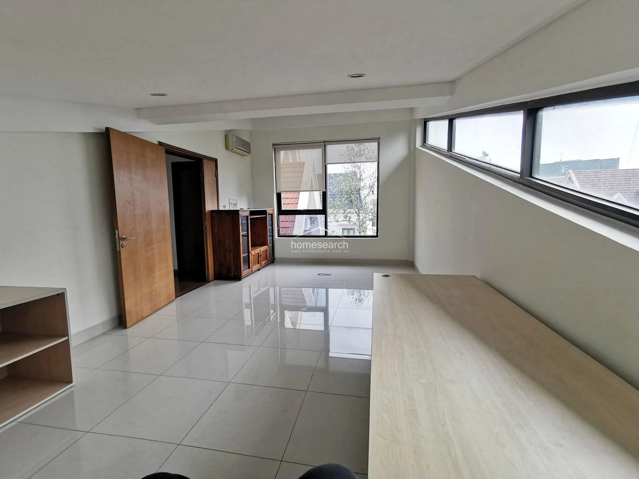 This functional home office or study area is characterized by a unique sloped ceiling and large, angled windows that offer ample natural light. The space includes a built-in wooden desk and cabinetry, polished tile floors, and a wall-mounted AC unit, presenting a well-maintained and professional environment.