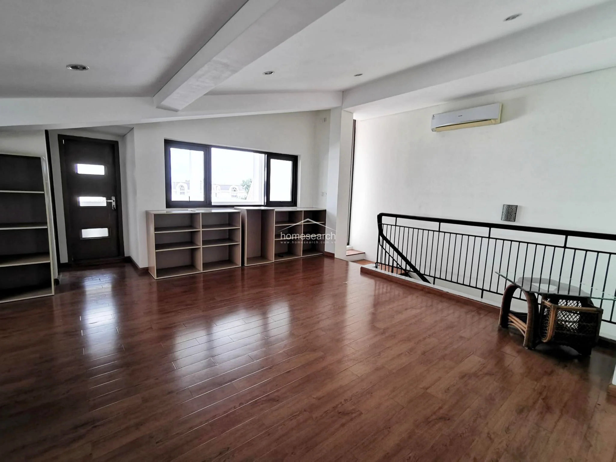 This spacious room features rich wood-laminate flooring and a sloped ceiling, creating an airy and open feel. It is equipped with a wall-mounted AC unit, extensive built-in open shelving for storage, and a dark wood door, maintaining a clean and orderly condition.