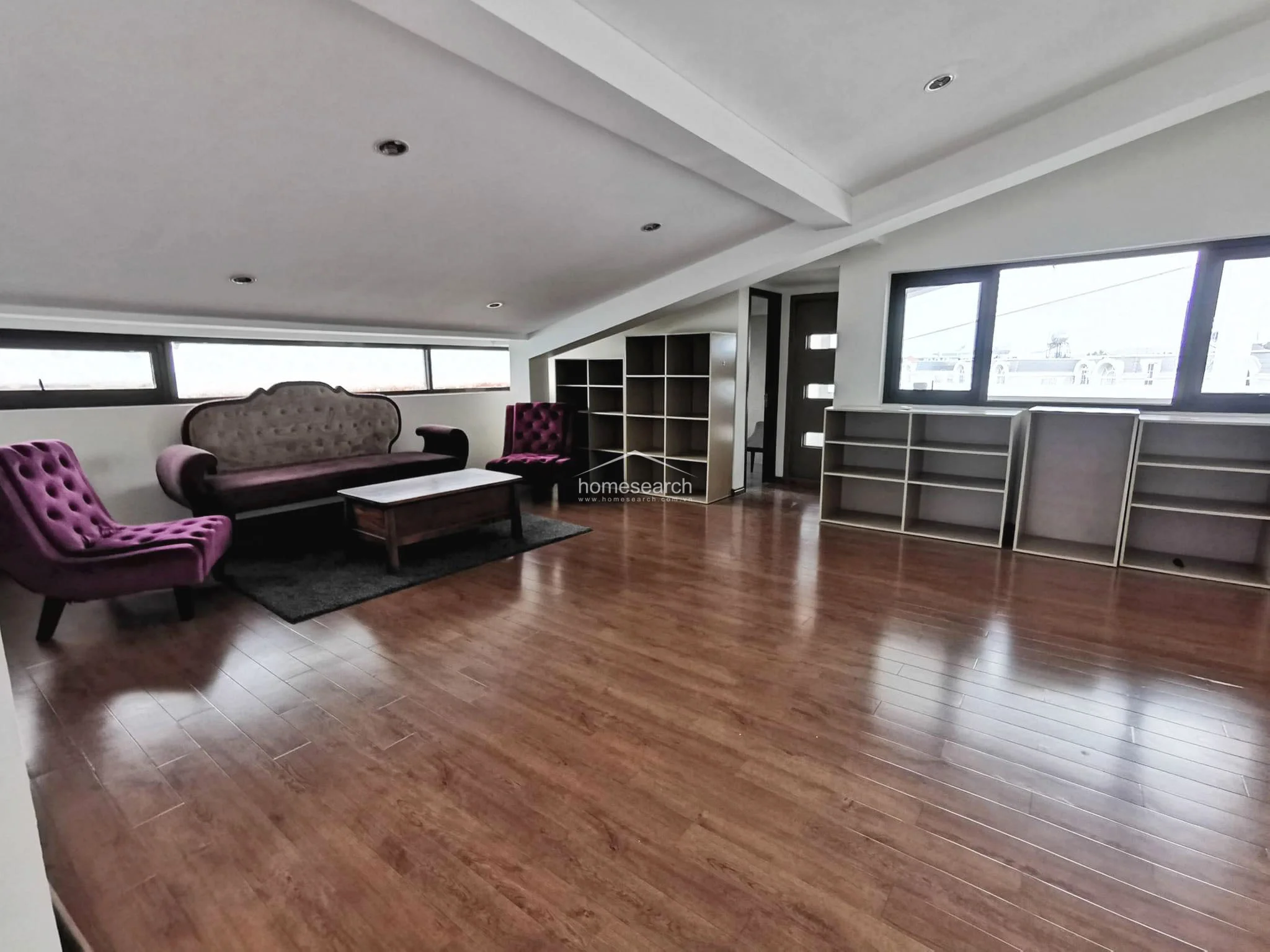This attic-style living area is furnished with elegant purple velvet seating, including a sofa and matching armchairs, arranged around a wooden coffee table. The room boasts warm wood-laminate flooring, multiple built-in storage units, and large windows that provide a bright, inviting atmosphere.