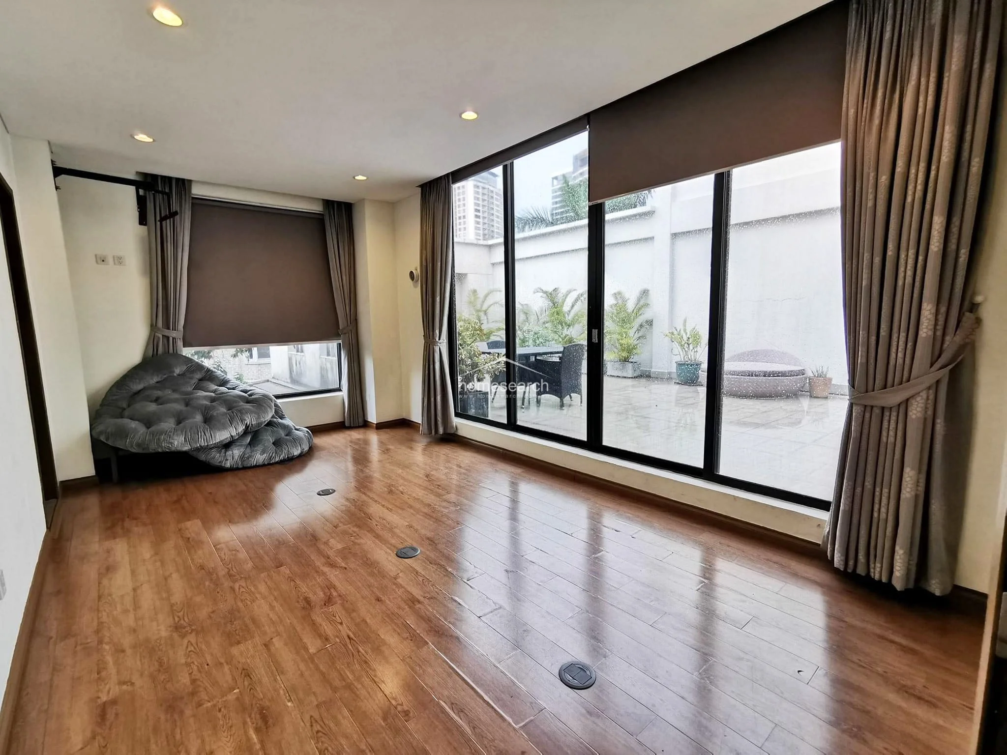 This living area features floor-to-ceiling glass doors that open onto a private terrace, allowing for excellent natural light and a seamless indoor-outdoor flow. The room is finished with warm wood flooring, neutral curtains, and a large, plush grey lounge chair, reflecting a modern and well-kept living space.