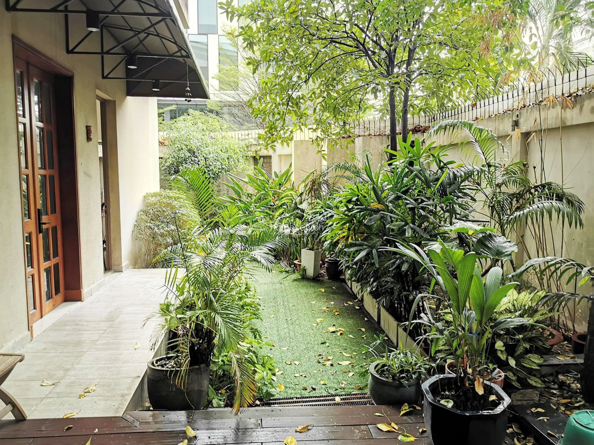This garden area serves as a lush, private outdoor retreat featuring a mix of artificial turf and a wooden deck transition. It is enclosed by high walls topped with security fencing, providing a secluded atmosphere filled with various potted tropical plants and a mature tree. The space is well-defined and offers a peaceful view from the adjacent living room.