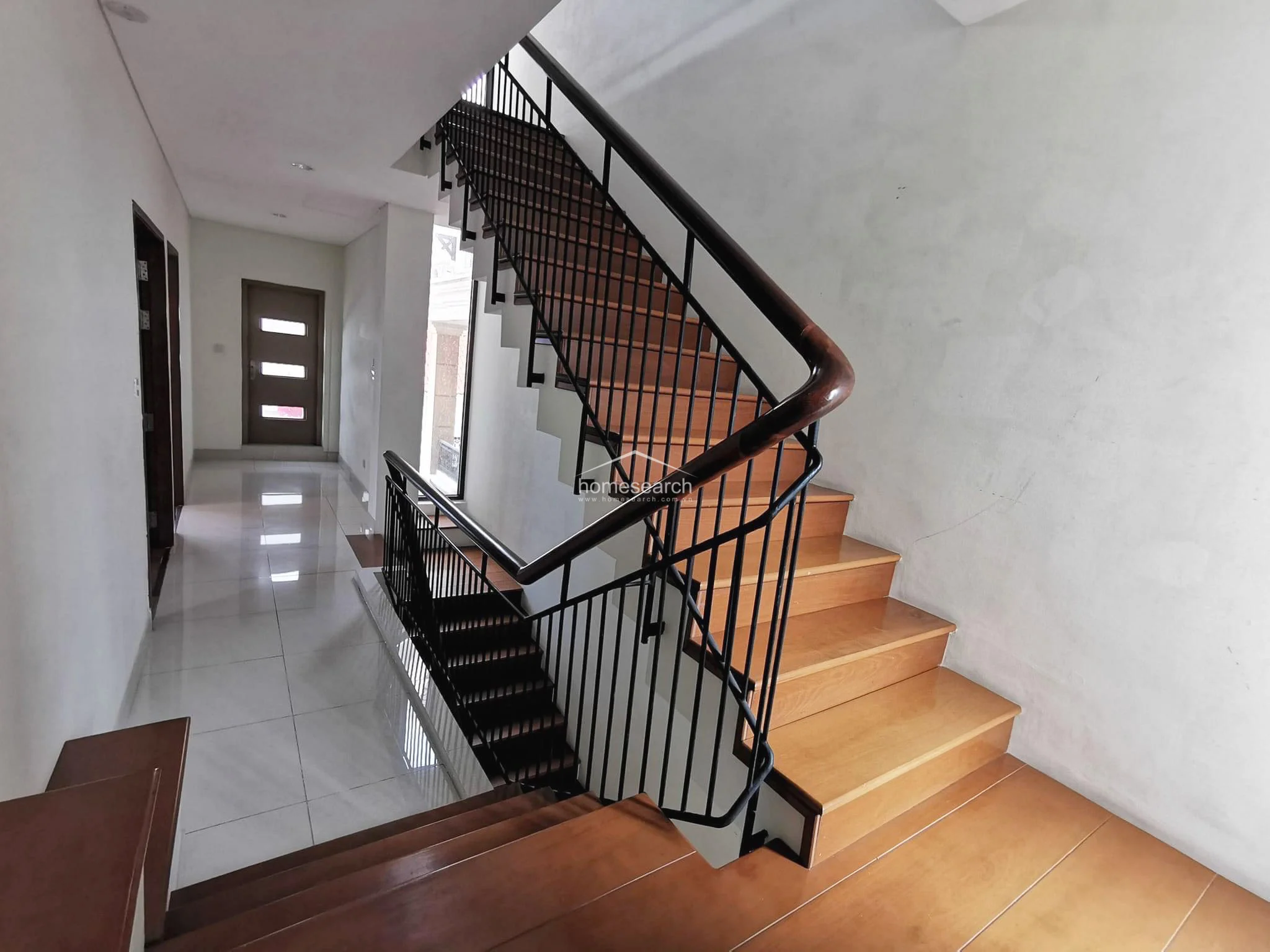 This architectural staircase serves as a central transition point, featuring wooden treads and a sleek black metal railing with a wooden handrail. The lower level is finished with light-colored floor tiles, contrasting with the warm wood of the stairs, while the walls remain a clean, neutral white. The design is modern and open, allowing for a clear visual flow between the different levels of the home.