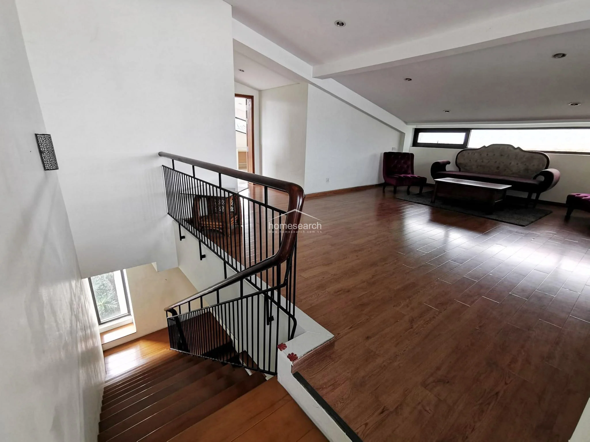 This upper-level landing area features warm wood-look flooring and a distinctive sloped ceiling, creating an open, airy feel. The space is anchored by a black metal railing with a wooden handrail and includes a vintage-style seating area illuminated by natural light from a horizontal window.