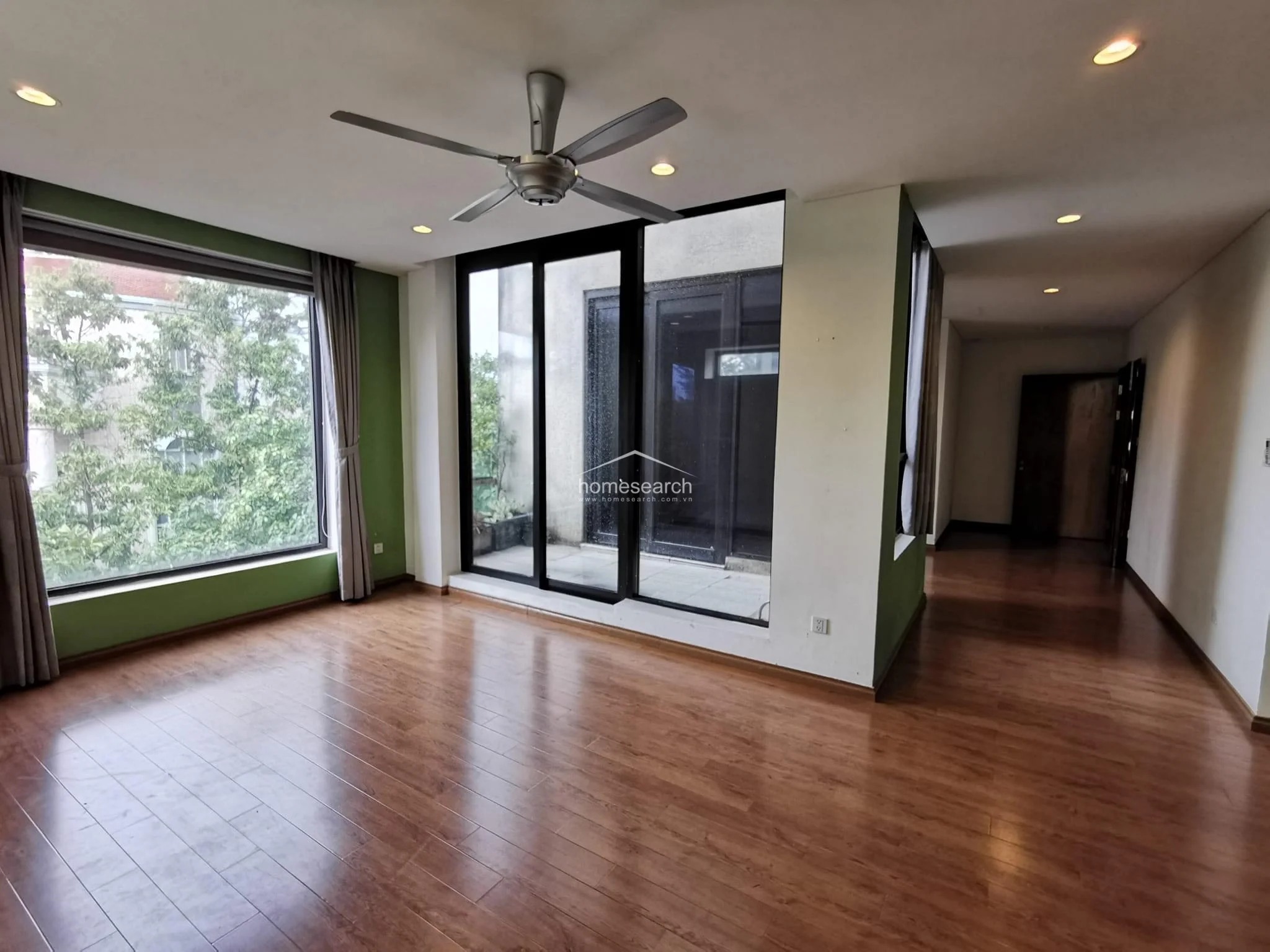 This bright living space features wood-look flooring, green accent walls, and a large window that offers a view of lush greenery. It is equipped with a modern ceiling fan and direct access to an outdoor balcony through sliding glass doors, enhancing the indoor-outdoor flow.